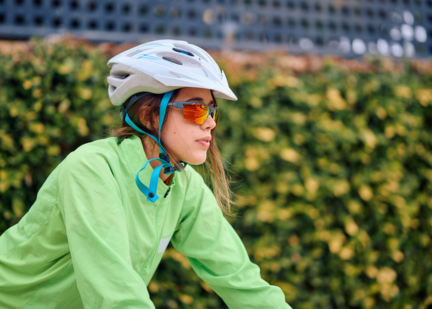 Radfahrerin mit Helm und Schutzbrille fährt vor grüner Hecke.